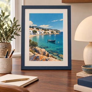 May include: A framed vertical art print featuring a coastal landscape with white buildings, a blue boat floating on clear turquoise water, and rocky foreground terrain under a bright blue sky. The frame is a dark navy blue with a white mat border.
