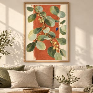 May include: A framed wall art print featuring a botanical illustration of a leafy branch with green foliage, small pale yellow berries, and a single blue berry, set against a vertical terracotta orange brushstroke on a cream background.