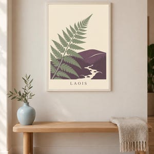 May include: A rectangular wall art print in a light-coloured frame featuring a stylised green fern frond overlapping a dark purple landscape with a winding white river and hills. The word LAOIS is printed in black capital letters at the bottom.