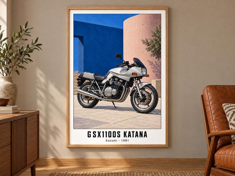 Puede incluir: Una l&aacute;mina enmarcada que muestra una motocicleta Suzuki GSX1100S Katana plateada aparcada sobre una superficie pavimentada. El fondo muestra una pared azul brillante y una estructura rosa curva. El texto GSX1100S KATANA Suzuki 1981 aparece debajo.