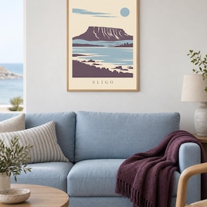 May include: A framed wall art print featuring a stylized landscape illustration of a mountain range near a body of water. The artwork uses a palette of cream, light blue, and dark purple, with the word SLIGO printed in black at the bottom center.