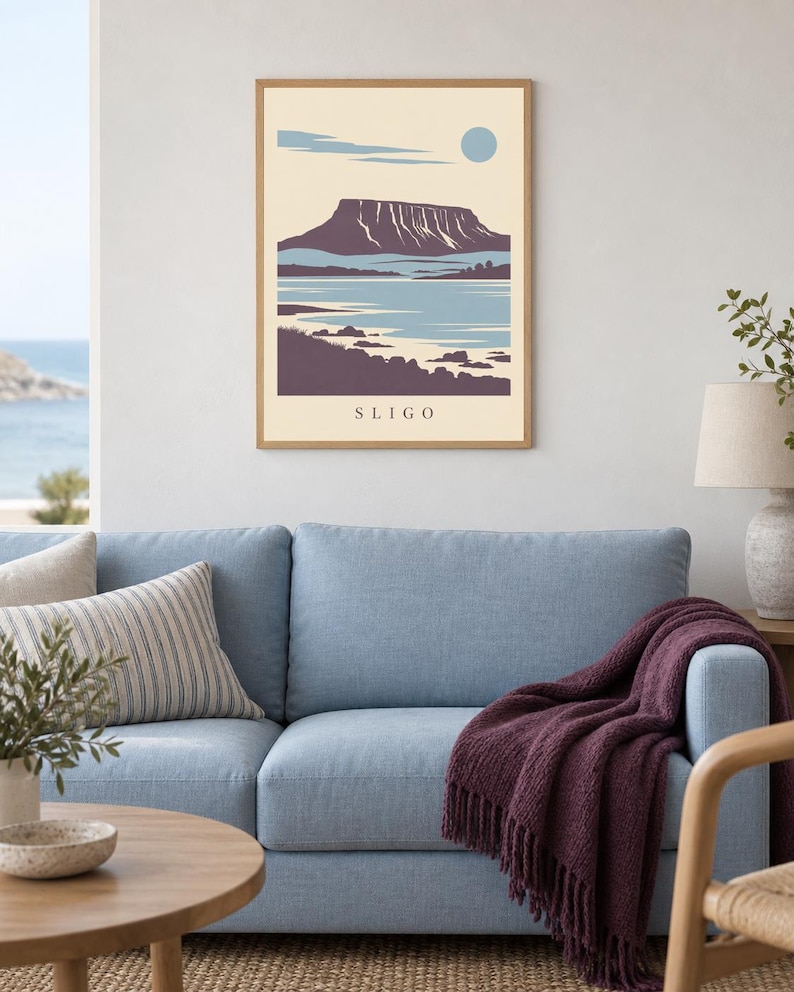 May include: A framed wall art print featuring a stylized landscape illustration of a mountain range near a body of water. The artwork uses a palette of cream, light blue, and dark purple, with the word SLIGO printed in black at the bottom center.