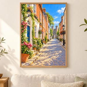 May include: A framed wall art print depicting a cobblestone street lined with brick buildings, vibrant pink and white flowers in terracotta pots, a blue door, and a stone archway in the distance under a bright blue sky with scattered white clouds.