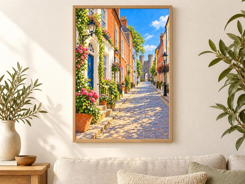 May include: A framed wall art print depicting a cobblestone street lined with brick buildings, vibrant pink and white flowers in terracotta pots, a blue door, and a stone archway in the distance under a bright blue sky with scattered white clouds.