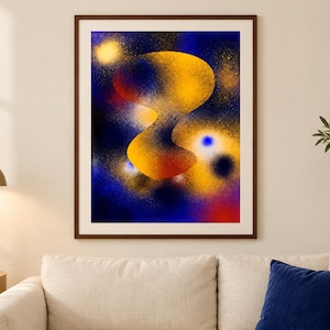 May include: A framed wall art print featuring an abstract composition with a central, organic yellow and orange shape set against a deep blue, spray-painted background. The artwork includes scattered golden speckles and glowing light effects.
