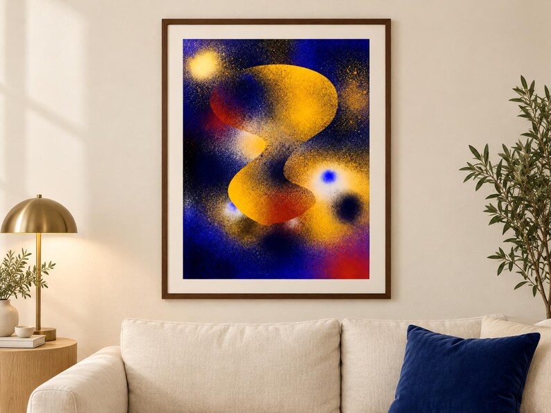 May include: A framed wall art print featuring an abstract composition with a central, organic yellow and orange shape set against a deep blue, spray-painted background. The artwork includes scattered golden speckles and glowing light effects.