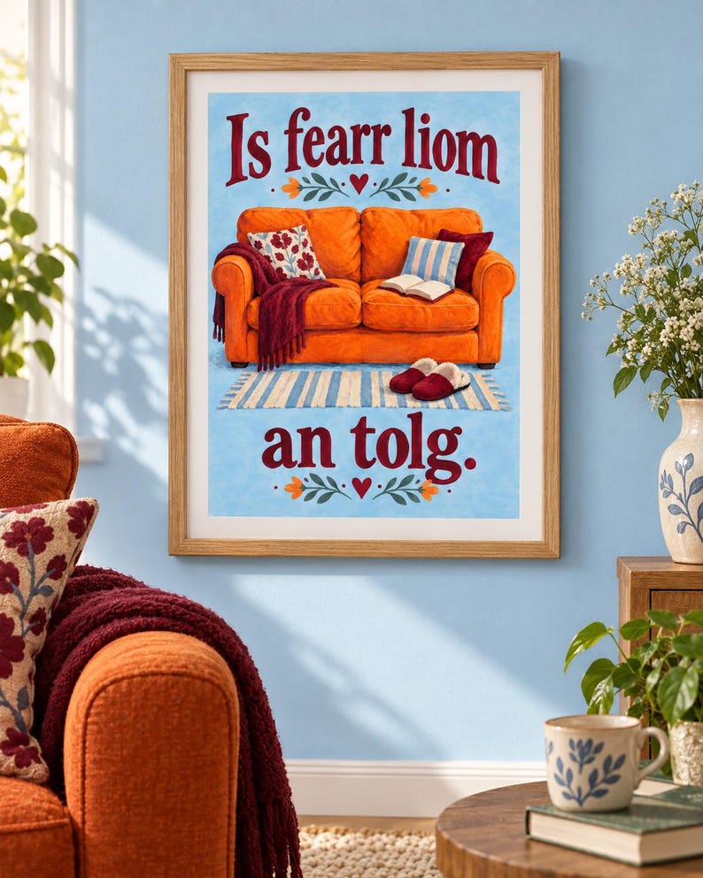 K&ouml;nnte beinhalten: Ein gerahmter Kunstdruck mit einer Illustration eines leuchtend orangefarbenen Sofas auf hellblauem Grund. Der Text "Is fearr liom an tolg" ist in dunkelroten Buchstaben geschrieben. Das Sofa hat eine weinrote Decke, Kissen und Hausschuhe.
