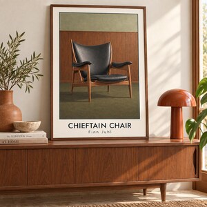 May include: A rectangular wall art print featuring a black leather armchair with a wooden frame. The text CHIEFTAIN CHAIR and Finn Juhl is printed in black at the bottom. The print is framed in a thin brown wood border and displayed on a surface.