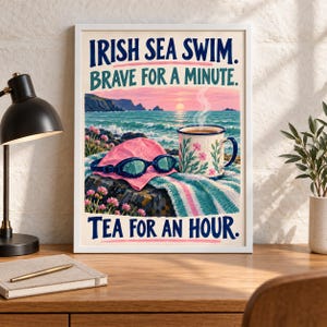 May include: A framed art print featuring a coastal scene with a pink swim cap, blue goggles, and a floral mug on a towel. The text reads: IRISH SEA SWIM. BRAVE FOR A MINUTE. TEA FOR AN HOUR. The background shows a sunset over the ocean and rocky cliffs.