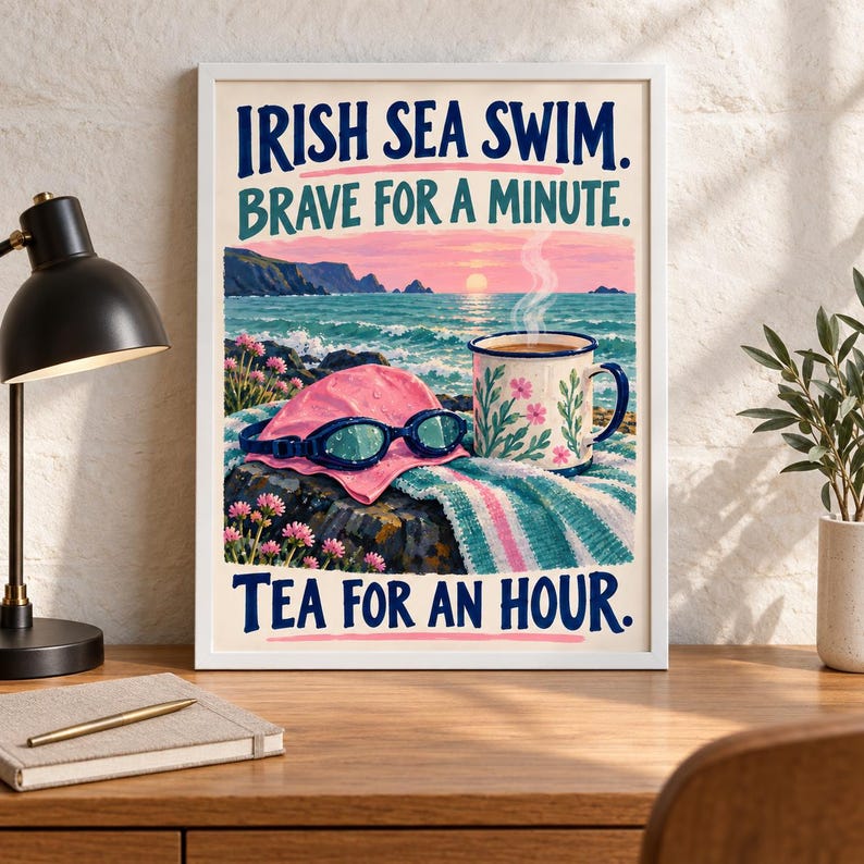 May include: A framed art print featuring a coastal scene with a pink swim cap, blue goggles, and a floral mug on a towel. The text reads: IRISH SEA SWIM. BRAVE FOR A MINUTE. TEA FOR AN HOUR. The background shows a sunset over the ocean and rocky cliffs.