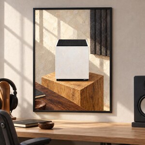 Op de afbeelding: Een ingelijste muurkunstprint met een witte kubusvormige luidspreker met een zwart bovenpaneel, rustend op een getextureerd houten blok. Het kunstwerk wordt getoond in een kamer met een houten bureau, een zwarte koptelefoon en een studiomonitor.