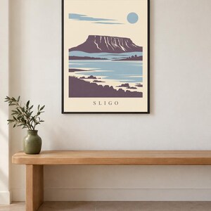 May include: A framed wall art print featuring a stylized landscape illustration of a flat-topped mountain, a body of water, and a pale blue sun against a cream background. The word SLIGO is printed in black capital letters at the bottom center.