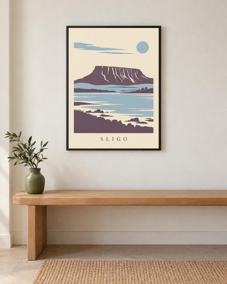 May include: A framed wall art print featuring a stylized landscape illustration of a flat-topped mountain, a body of water, and a pale blue sun against a cream background. The word SLIGO is printed in black capital letters at the bottom center.