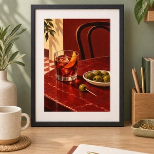 May include: A framed art print depicting a glass of dark red cocktail with ice cubes and an orange peel garnish, placed on a deep red table next to a small white bowl filled with green olives and a single olive on a wooden toothpick.