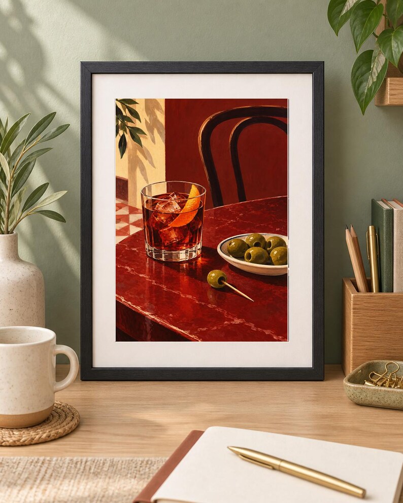 May include: A framed art print depicting a glass of dark red cocktail with ice cubes and an orange peel garnish, placed on a deep red table next to a small white bowl filled with green olives and a single olive on a wooden toothpick.