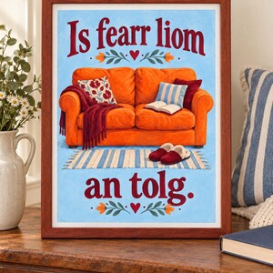 K&ouml;nnte beinhalten: Ein gerahmter Druck mit einer Illustration eines leuchtend orangefarbenen Sofas auf hellblauem Grund. Der Text 'Is fearr liom an tolg.' ist in Rot geschrieben. Das Sofa hat eine weinrote Decke, gemusterte Kissen, ein offenes Buch und Hausschuhe.
