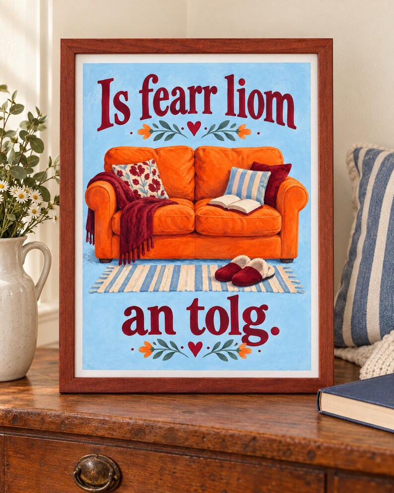 K&ouml;nnte beinhalten: Ein gerahmter Druck mit einer Illustration eines leuchtend orangefarbenen Sofas auf hellblauem Grund. Der Text 'Is fearr liom an tolg.' ist in Rot geschrieben. Das Sofa hat eine weinrote Decke, gemusterte Kissen, ein offenes Buch und Hausschuhe.