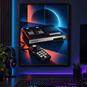 Op de afbeelding: Een ingelijste wandkunstprint met een zwarte ColecoVision-spelconsole en een bijbehorende bedrade controller. De console toont de merknaam en heeft een zilverkleurig voorpaneel. De achtergrond toont een gestileerde blauwe en oranje cirkelvormige afbeelding.