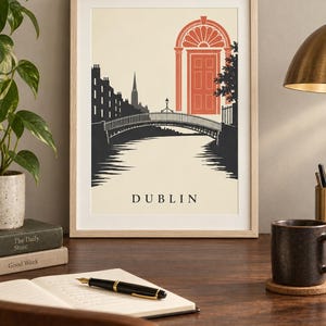 K&ouml;nnte beinhalten: Ein gerahmter Kunstdruck mit einer stilisierten Illustration von Dublin, Irland. Die Grafik zeigt eine dunkelgraue Bogenbr&uuml;cke &uuml;ber einem Fluss, eine Stadtsilhouette und eine markante korallenfarbene georgianische T&uuml;r. Das Wort DUBLIN steht unten.