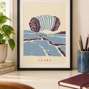 May include: A framed art print featuring a stylized illustration of a concertina floating above a blue and purple landscape of cracked ground. The word CLARE is printed in black capital letters at the bottom center of the cream-colored background.