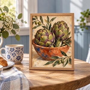 May include: A framed wall art print featuring a painting of two green and purple artichokes resting in a rustic, weathered terracotta bowl, accompanied by olive branches with small green olives, set against a light, textured cream background.