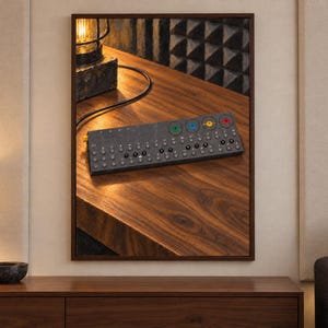 Op de afbeelding: Een donkergrijze draagbare synthesizer met een raster van kleine ronde knoppen, een rij genummerde toetsen en vier opvallende draaiknoppen in groen, blauw, geel en rood, geplaatst op een houten oppervlak naast een verlichte bureaulamp.