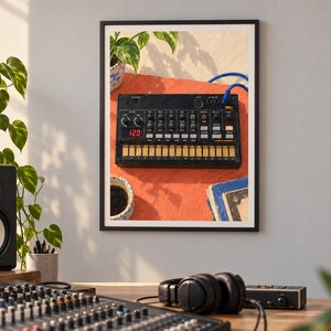Puede incluir: Una impresi&oacute;n de pared enmarcada que muestra un sintetizador Korg Volca negro con una pantalla digital roja que indica 120, colocado sobre una superficie naranja junto a un cable azul, una taza de cer&aacute;mica peque&ntilde;a y libros con cubiertas azules.