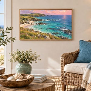 May include: A rectangular landscape painting in a light wood frame depicting a coastal scene with turquoise water, rocky cliffs, and green grassy hills under a pink and orange sunset sky, displayed on a wall above a wicker chair and wooden table.