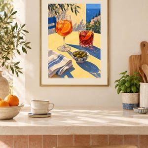 May include: A framed art print depicting a yellow table with an orange cocktail in a stemmed glass, a dark red cocktail in a tumbler, and a small bowl of green olives. A blue and white striped napkin sits on the table, with a coastal view in the background.