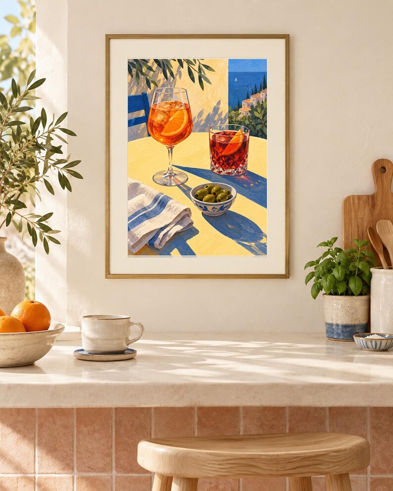 May include: A framed art print depicting a yellow table with an orange cocktail in a stemmed glass, a dark red cocktail in a tumbler, and a small bowl of green olives. A blue and white striped napkin sits on the table, with a coastal view in the background.