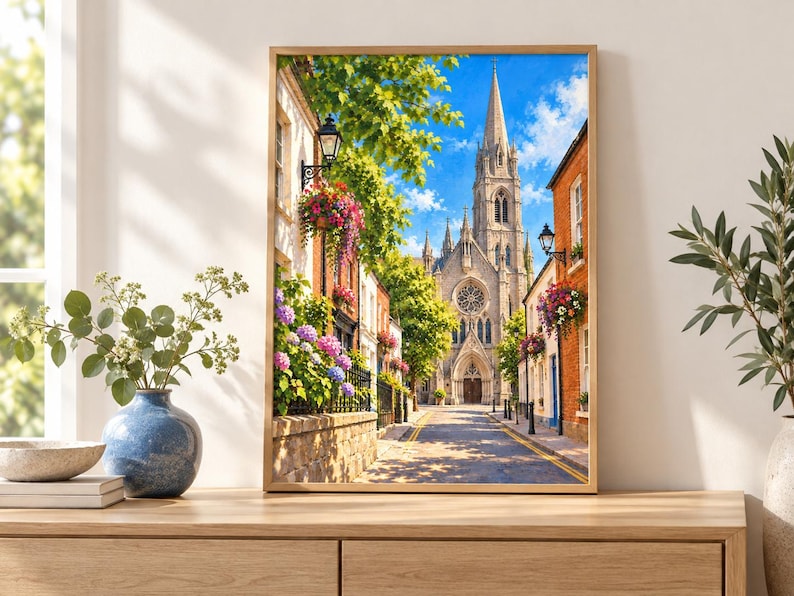Pu&ograve; includere: Una stampa artistica incorniciata che raffigura una soleggiata strada europea con un'alta chiesa in pietra dotata di guglia e rosone, fiancheggiata da edifici in mattoni, cesti di fiori appesi e vivaci cespugli di ortensie blu, rosa e viola.