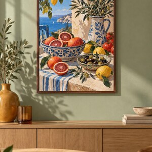 Op de afbeelding: Een ingelijste kunstdruk met een mediterrane sc&egrave;ne met een blauw-wit patroon kom gevuld met bloedsinaasappels, een bijpassende kan, een kom met olijven en citroenen op een tafel bedekt met een blauw-wit gestreept kleed.