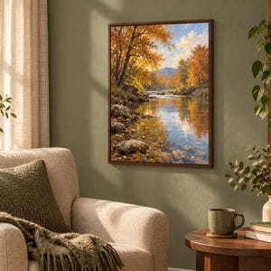 K&ouml;nnte beinhalten: Ein rechteckiges gerahmtes Wandbild, das eine herbstliche Landschaft mit einem flie&szlig;enden Fluss, felsigen Ufern und leuchtend gold-orangen B&auml;umen unter einem blauen Himmel mit wei&szlig;en Wolken zeigt, montiert an einer olivgr&uuml;nen Wand &uuml;ber einem beigen Sessel.