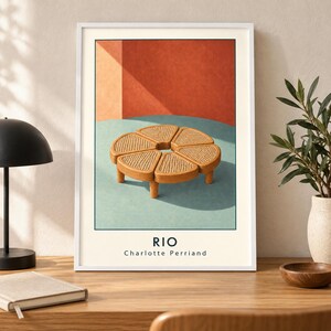 May include: A framed art print featuring a circular wooden coffee table with six wedge-shaped sections and woven cane inserts. The table sits on a light blue surface against a terracotta wall. The text RIO and Charlotte Perriand is printed below.
