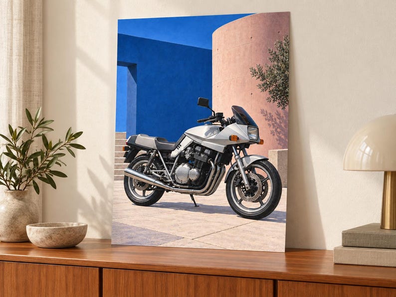 Puede incluir: Una motocicleta plateada estacionada sobre una superficie pavimentada frente a una pared azul brillante y una estructura curva de color melocot&oacute;n. La motocicleta cuenta con un asiento negro, un parabrisas oscuro y componentes mec&aacute;nicos del motor expuestos con tubos de escape cromados.
