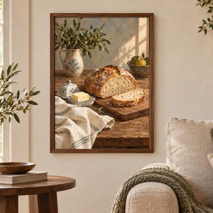 Peut inclure: Une impression d'art encadr&eacute;e repr&eacute;sentant une nature morte rustique sur une table en bois. L'image montre une miche de pain au levain ronde, une tranche de pain, un beurrier en c&eacute;ramique avec une noisette de beurre jaune, un pichet et un bol.