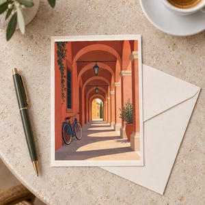 Pu&ograve; includere: Un biglietto d'auguri rettangolare con l'illustrazione di un porticato color terracotta, una bicicletta blu appoggiata a una parete, lanterne sospese e una pianta in vaso, accompagnato da una busta color crema e una penna scura.