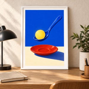 May include: A framed art print featuring a vibrant blue spoon hovering above a red saucer. A bright yellow sphere rests inside the spoon. The background is divided into a solid blue upper section and a light cream lower section with shadows.