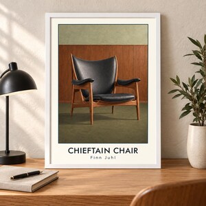 May include: A framed wall art print featuring a black leather armchair with a wooden frame. The text CHIEFTAIN CHAIR and Finn Juhl is printed in black at the bottom. The print is displayed in a white frame on a wooden desk surface.