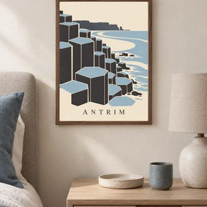 May include: A framed wall art print featuring a stylised illustration of the Giant's Causeway in Antrim. The graphic depicts dark grey hexagonal basalt columns against a light blue sea and cream background, with the text ANTRIM printed at the bottom.