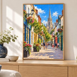 Op de afbeelding: Een ingelijste wandkunstprint met een zonnig Europees straatbeeld met kleurrijke winkelpuien, hangende bloemenmanden en voetgangers die naar een hoge stenen kerktoren lopen onder een helderblauwe lucht met witte wolken.