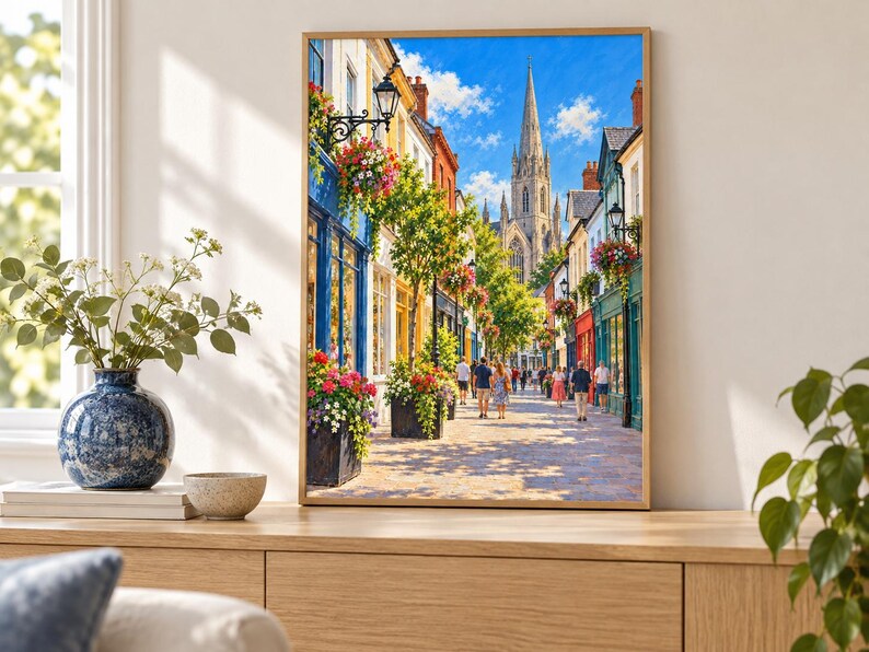Op de afbeelding: Een ingelijste wandkunstprint met een zonnig Europees straatbeeld met kleurrijke winkelpuien, hangende bloemenmanden en voetgangers die naar een hoge stenen kerktoren lopen onder een helderblauwe lucht met witte wolken.