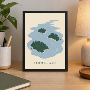 May include: A framed art print featuring a stylized illustration of a blue lake with three green islands covered in trees and a small white boat leaving a wake. The word FERMANAGH is printed in black capital letters at the bottom of the print.