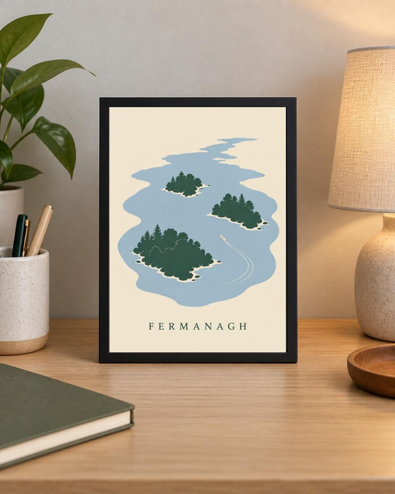 May include: A framed art print featuring a stylized illustration of a blue lake with three green islands covered in trees and a small white boat leaving a wake. The word FERMANAGH is printed in black capital letters at the bottom of the print.