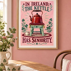 May include: A framed wall art print featuring a red kettle resting on a small stool, surrounded by green clover illustrations on a pink background. The text reads: IN IRELAND, THE KETTLE HAS SENIORITY. A small white cup sits on a rug below the stool.