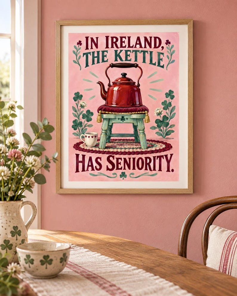 May include: A framed wall art print featuring a red kettle resting on a small stool, surrounded by green clover illustrations on a pink background. The text reads: IN IRELAND, THE KETTLE HAS SENIORITY. A small white cup sits on a rug below the stool.