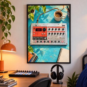 Puede incluir: Una impresi&oacute;n de arte de pared enmarcada que muestra una caja de ritmos Korg Electribe ER-1 sobre una superficie azul texturizada, acompa&ntilde;ada de una taza de caf&eacute;, un cuaderno y una planta, exhibida en una habitaci&oacute;n con escritorio, l&aacute;mpara y auriculares.