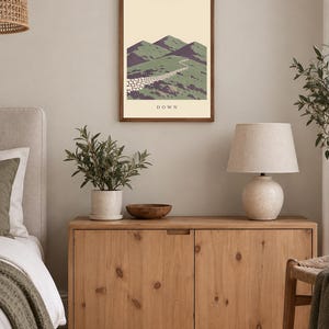 Pu&ograve; includere: Una stampa artistica da parete incorniciata che presenta un'illustrazione paesaggistica stilizzata di montagne verdi e un muro di pietra che si snoda tra le colline. L'opera ha uno sfondo color crema e la parola DOWN stampata in nero al centro in basso.