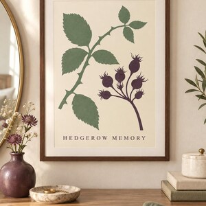 May include: A framed wall art print featuring a botanical illustration of green leaves and dark purple rose hips on a cream background. The text HEDGEROW MEMORY is printed in black at the bottom center of the print, which is enclosed in a dark wood frame.