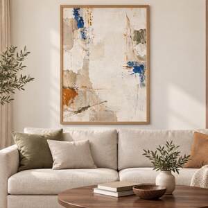 K&ouml;nnte beinhalten: Ein rechteckiges Wandbild in einem hellen Holzrahmen mit einer abstrakten Komposition aus strukturierten Schichten in Creme, Wei&szlig; und Beige, akzentuiert durch kr&auml;ftige Farbspritzer in Dunkelblau und gebranntem Orange.